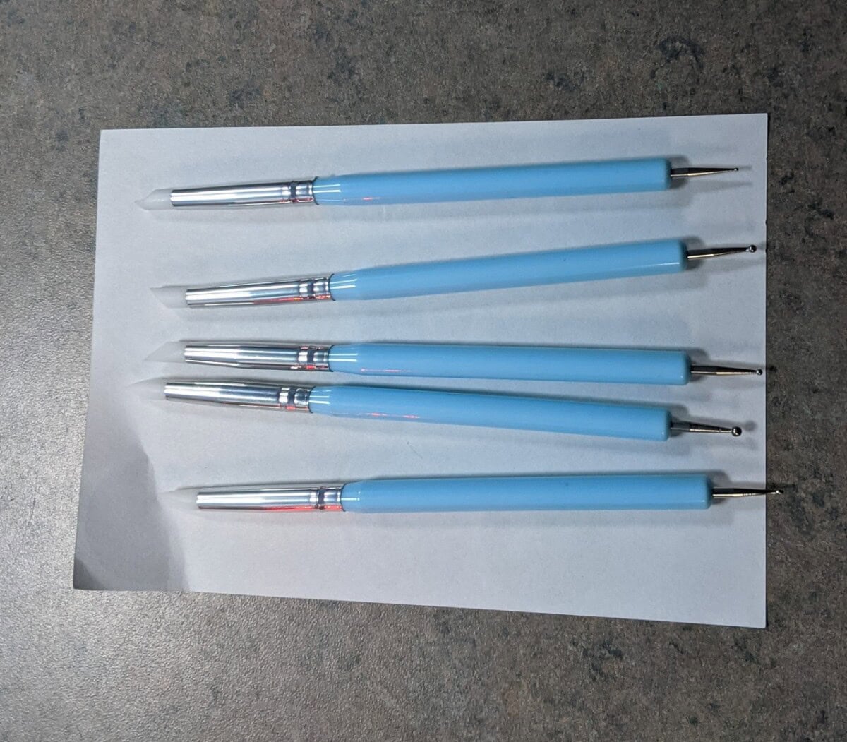 Five blue-handled dotting tools with metal tips are arranged side by side on a sheet of white paper, resting on a gray textured surface.