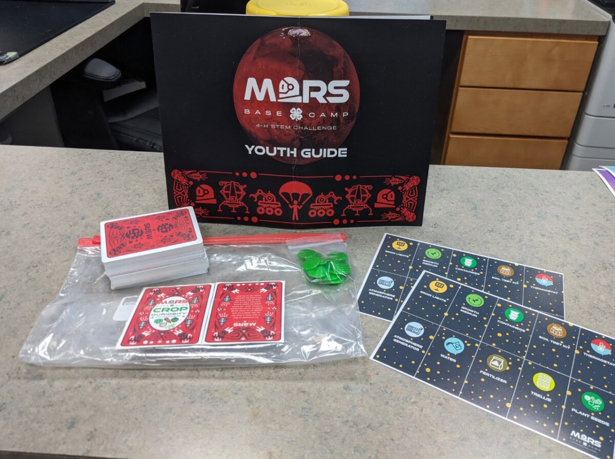 A Mars Base Camp Youth Guide display sits on a counter with a stack of playing cards, two green game pieces, and boards with colorful planets and icons arranged nearby.