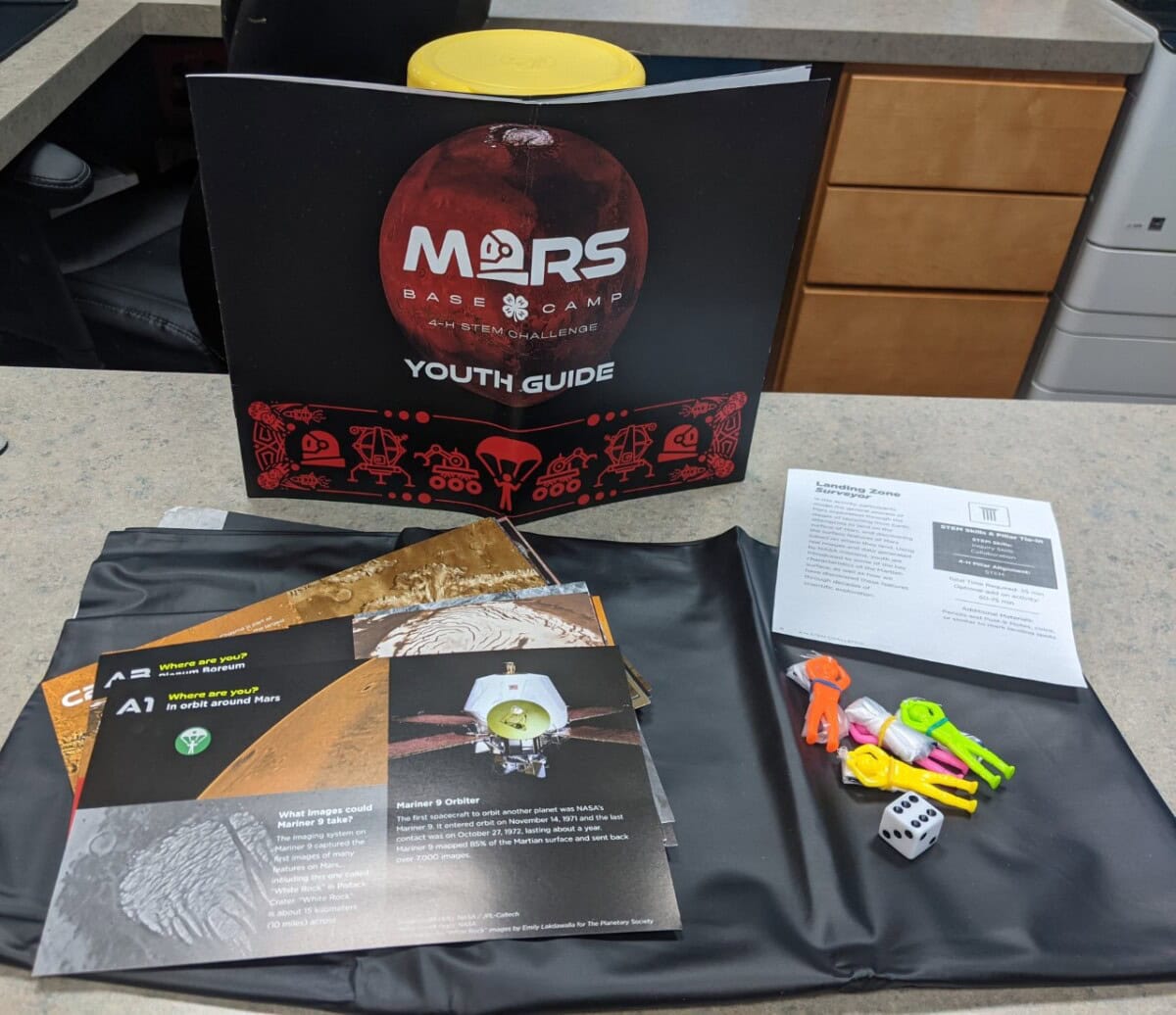 A STEM activity kit titled Mars Base Camp Youth Guide is displayed on a table with instruction cards, a folder, plastic game pieces, a die, and a yellow container. The kit has a Mars exploration theme.