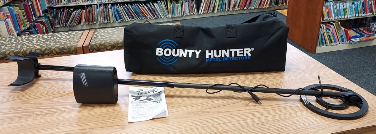 A Bounty Hunter metal detector is displayed on a wooden table in a library, with its carrying bag and instruction manual nearby. Bookshelves filled with books are visible in the background.