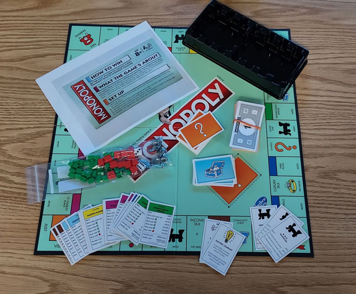 Monopoly board game set up on a wooden table with the board, instruction sheet, cards, money, houses, hotels, dice, and playing tokens arranged in the center. The box insert is placed to the side.