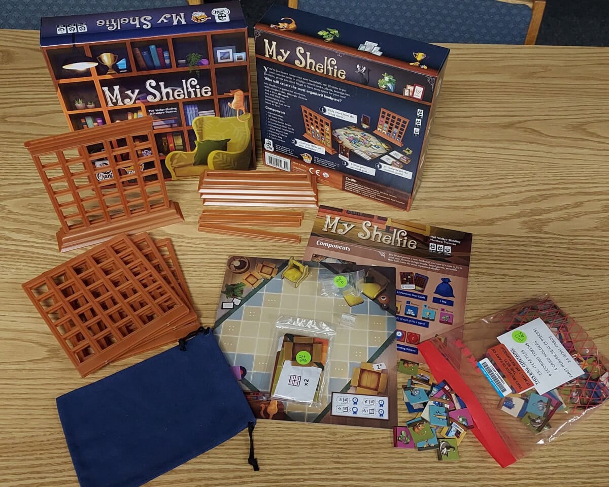 Board game components for My Shelfie are spread out on a wooden table, including a game box, plastic shelf frames, cardboard tokens, a cloth bag, instruction booklet, and game board.