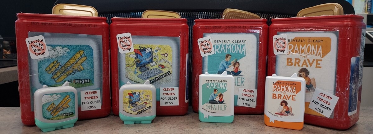 Five red audiobook cases and three compact audiobook players for children are displayed on a counter. The titles include Flight, Astronauts, Ramona, and Ramona the Brave, by Beverly Cleary.