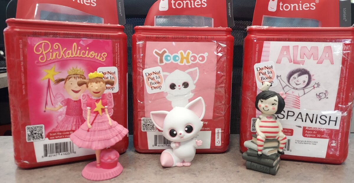 Three red Tonie audiobook boxes are displayed side by side. In front of each box is a small figurine: a pink princess, a white wide-eyed animal, and a girl sitting on books. Each box features colorful book cover art.