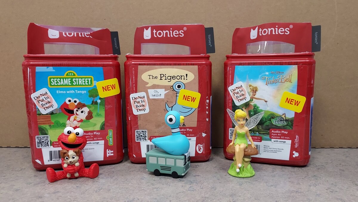 Three red Tonies audio boxes are side by side, featuring Sesame Street, The Pigeon, and Disney’s Tinker Bell. Figurines of Elmo, The Pigeon, and Tinker Bell are placed in front of their respective boxes.