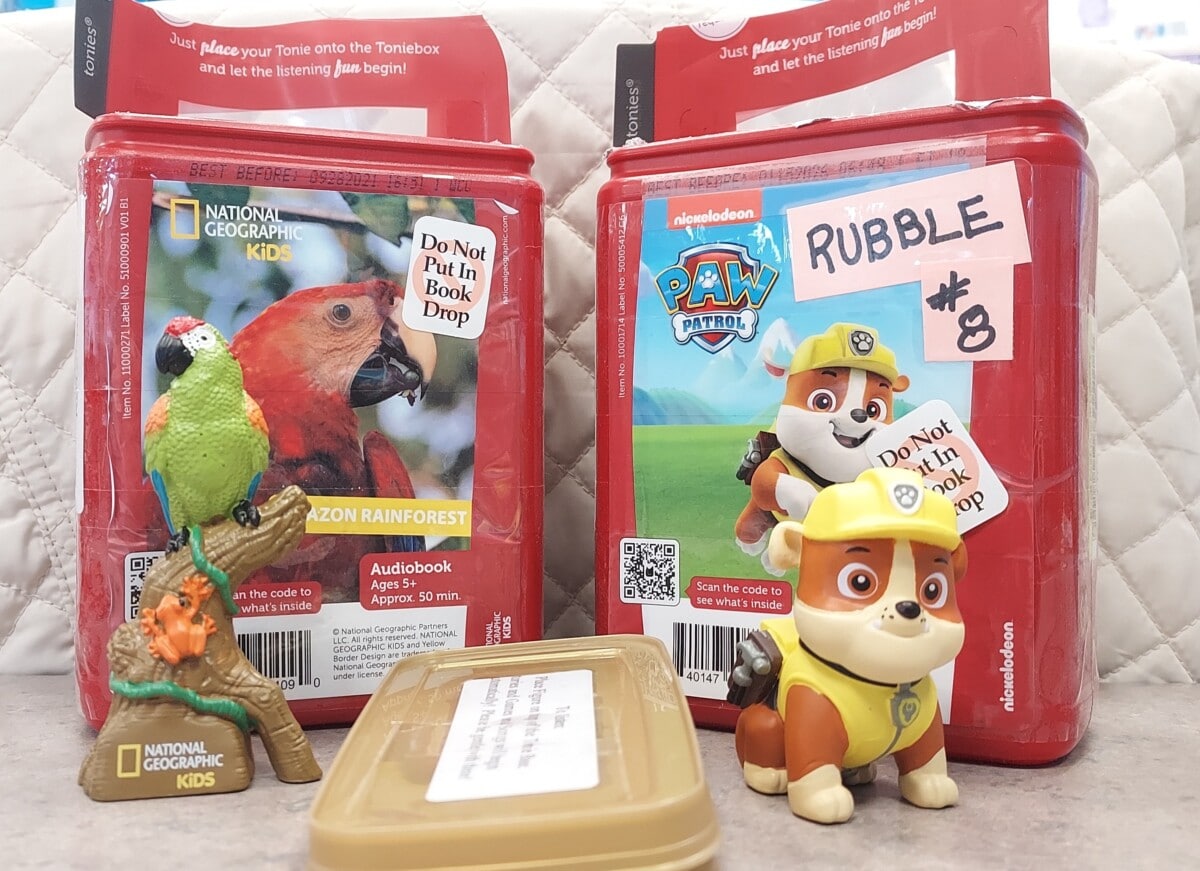 Two Toniebox audio characters: one featuring a parrot with a rainforest audiobook, and another with Paw Patrol character Rubble. Both have Do Not Put in Book Drop labels. Toy figures of a parrot and Rubble are in front.