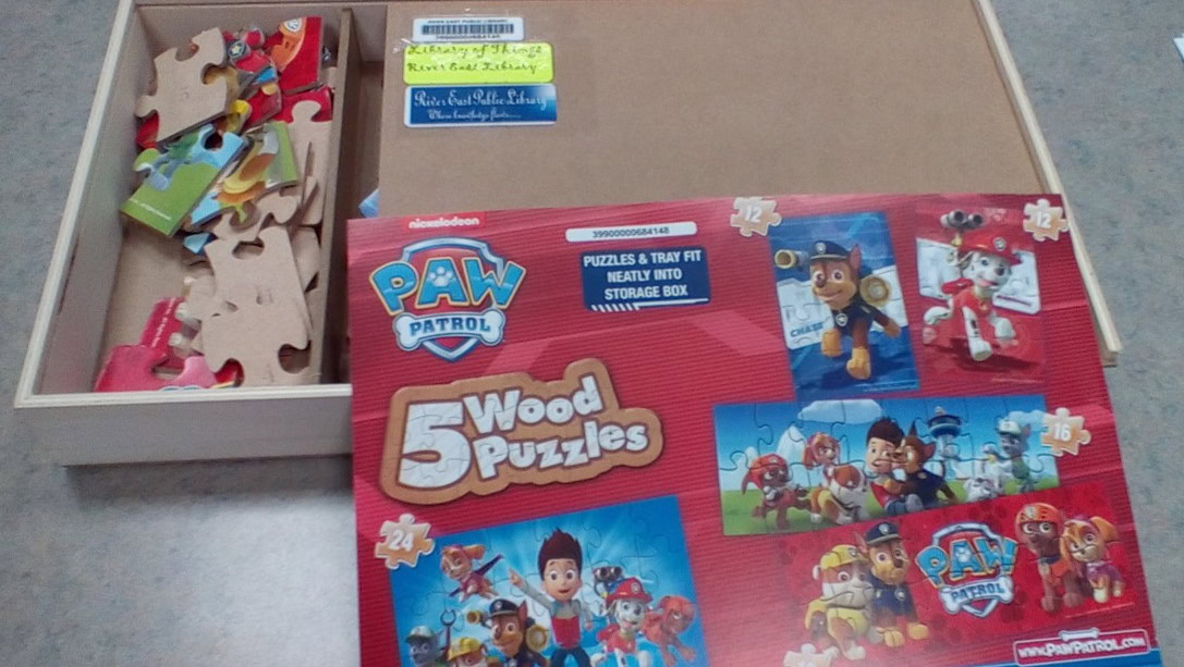 A wooden box with PAW Patrol-themed wooden puzzles, some puzzle pieces are inside the box. The puzzle cover shows images of PAW Patrol characters and mentions 5 Wood Puzzles with 24 and 32 pieces each.