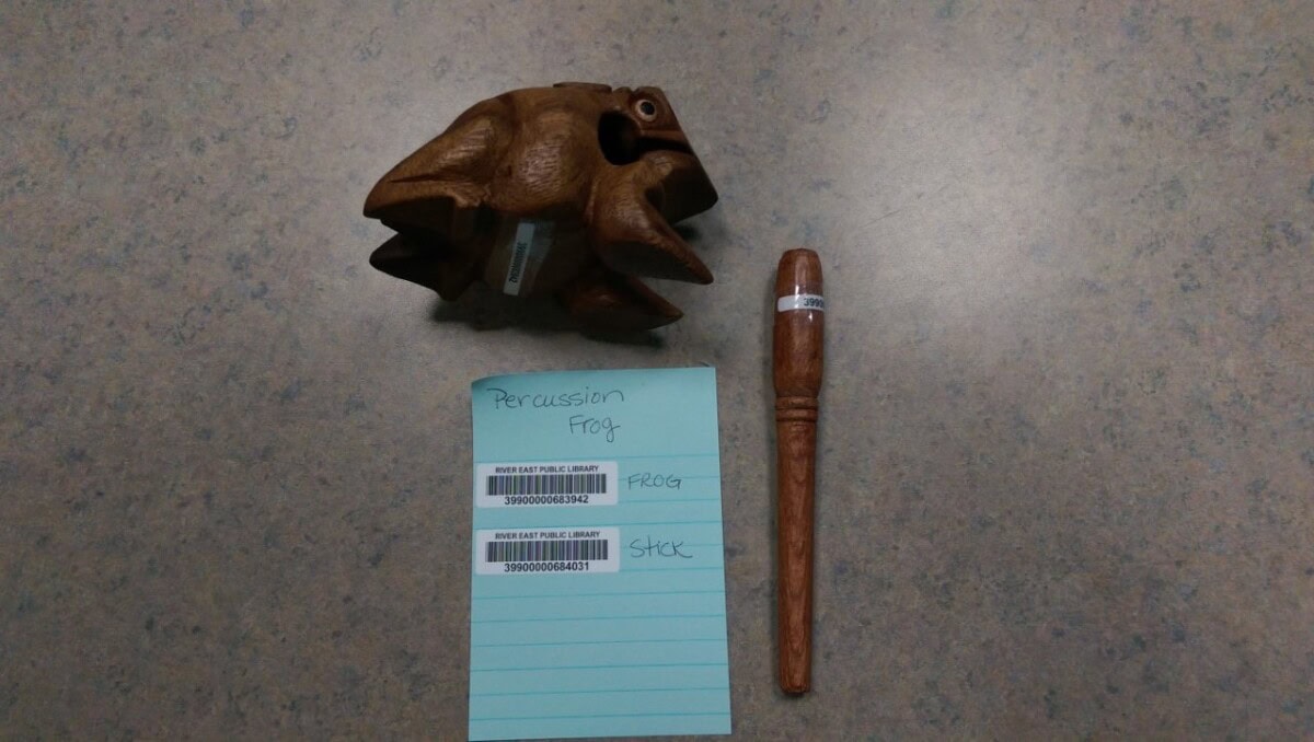 A wooden percussion frog and stick are on a gray table next to a blue note labeled Percussion Frog with barcodes and ID numbers. The frog and stick both have barcode labels attached.