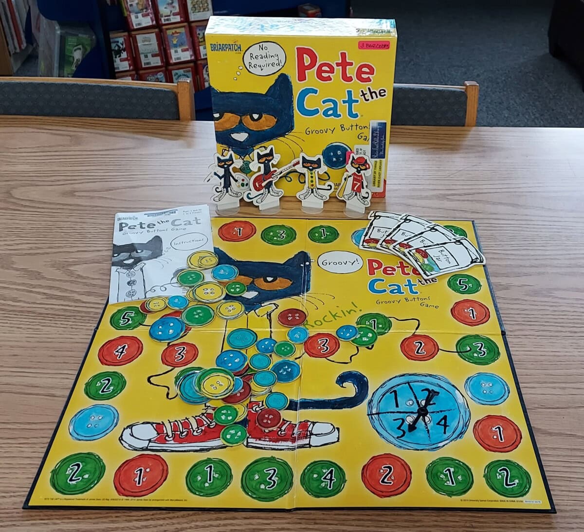 The board game Pete the Cat: The Groovy Buttons is set up on a wooden table, showing the colorful game board, character pieces, cards, a spinner, and the game box in the background.
