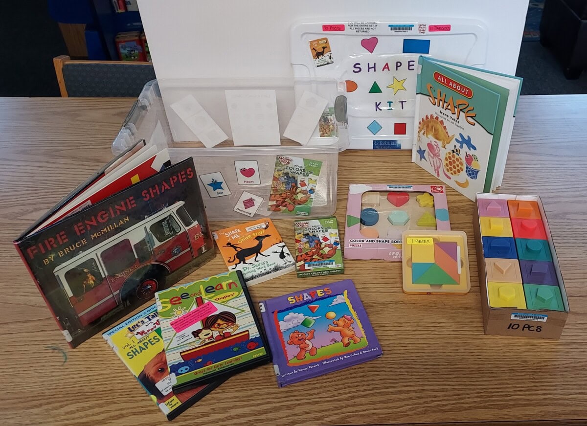 A tabletop display of children’s books, puzzles, games, and learning cards about shapes, including books titled “Fire Engine Shapes” and “All About Shapes.” A clear box labeled “Shape Kit” holds the items.