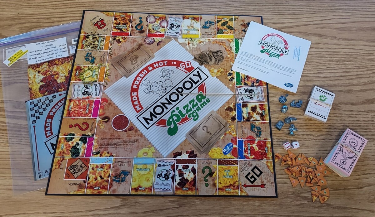Monopoly Pizza Game is displayed on a wooden table with its board, cards, dice, pizza-slice tokens, instruction sheet, and a zippered bag for pieces. The board features pizza-themed spaces and colorful artwork.