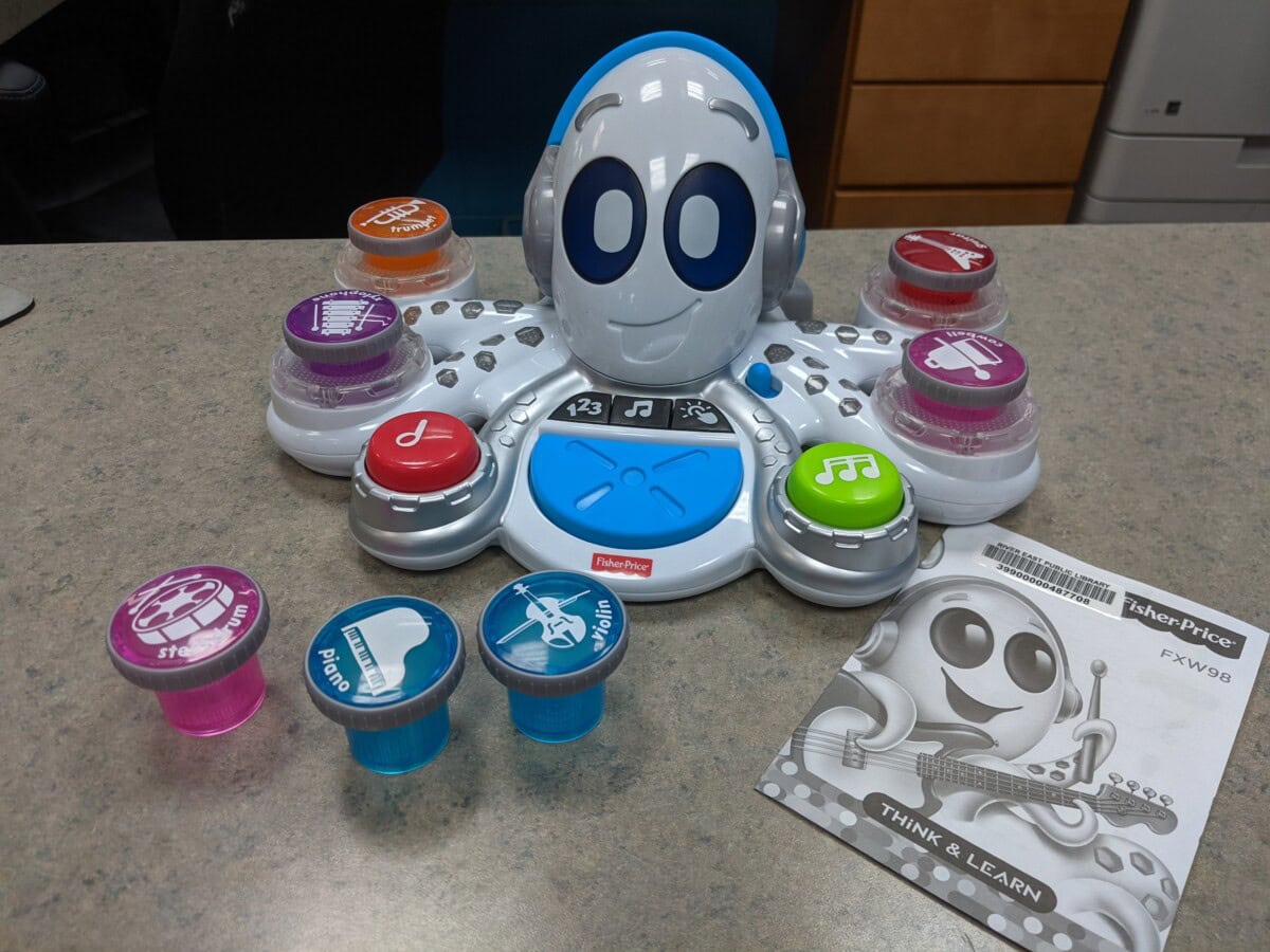 A Fisher-Price Think & Learn music toy shaped like a robot with various colorful buttons and removable discs on a countertop, alongside an instruction manual.