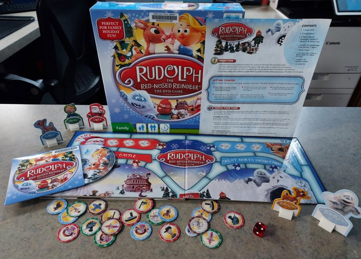 The image shows the board game Rudolph the Red-Nosed Reindeer: The DVD Game with its box, instructions, game board, character pieces, and various tokens arranged on a tabletop.