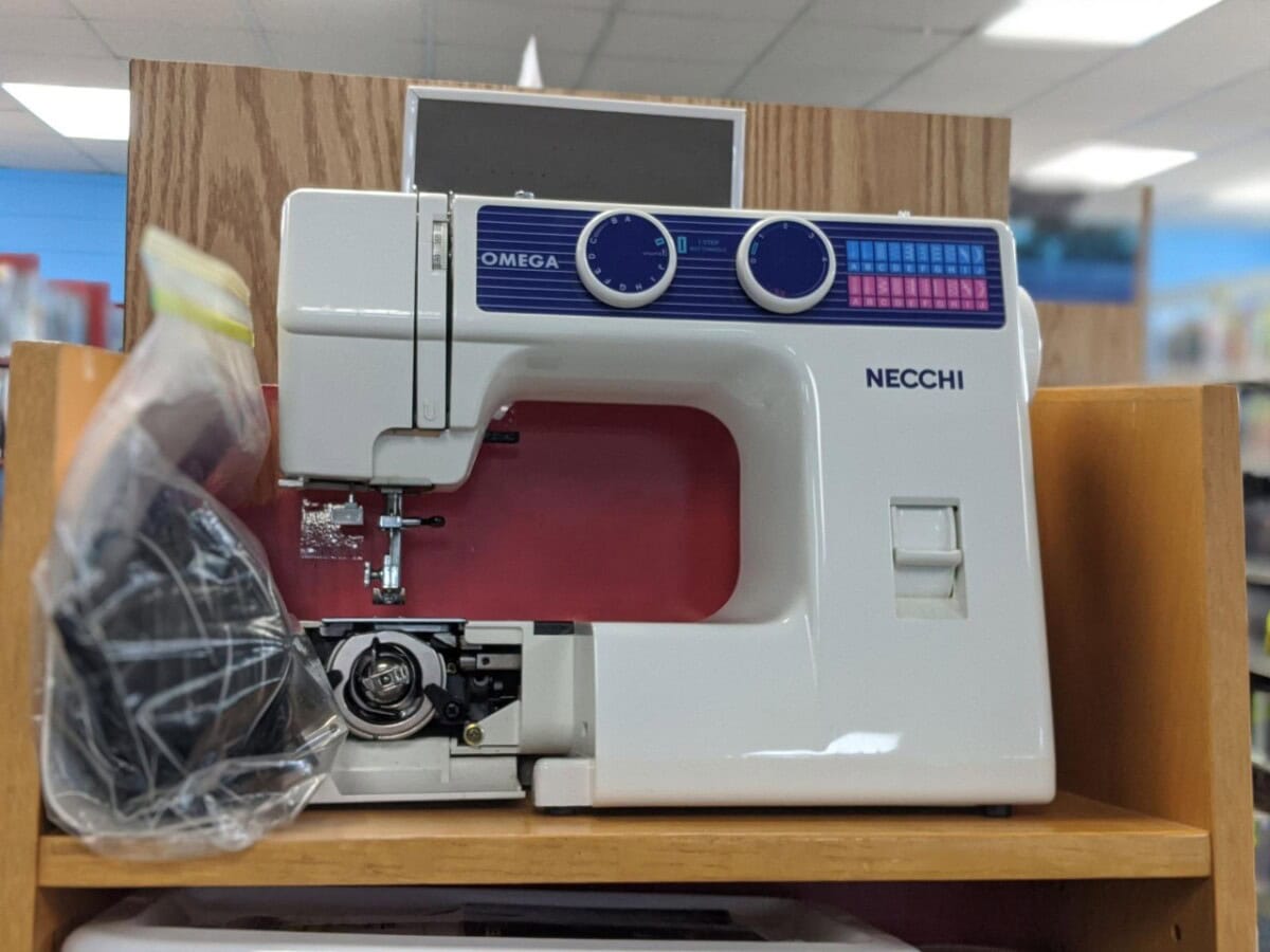 A Necchi Omega sewing machine sits on a wooden shelf, partially open to reveal its bobbin area. A plastic bag containing accessories hangs on the side. The machine is white with blue and pink accents.