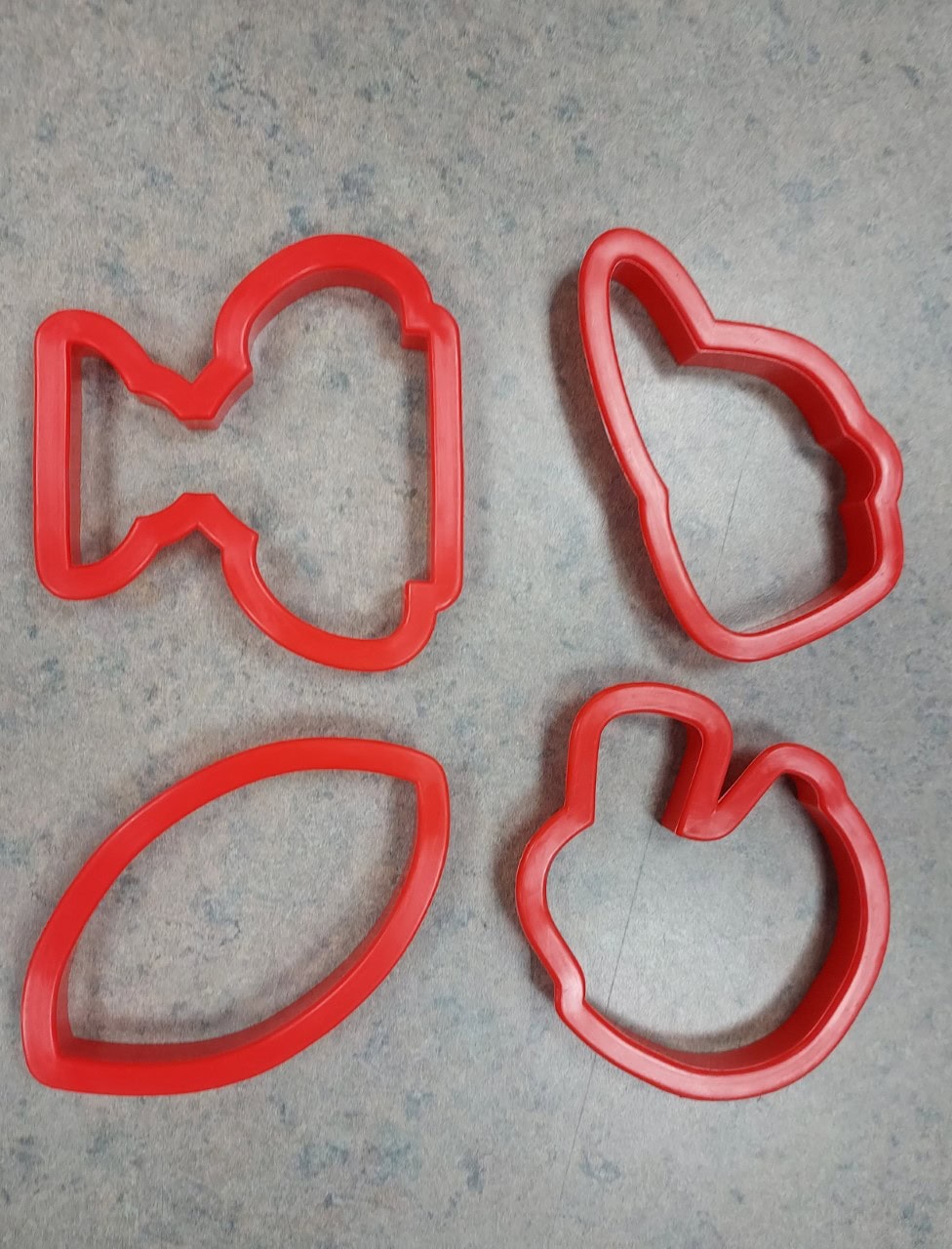Four red cookie cutters on a gray surface; shapes include a fish, a bunny head, a football, and a mitten.