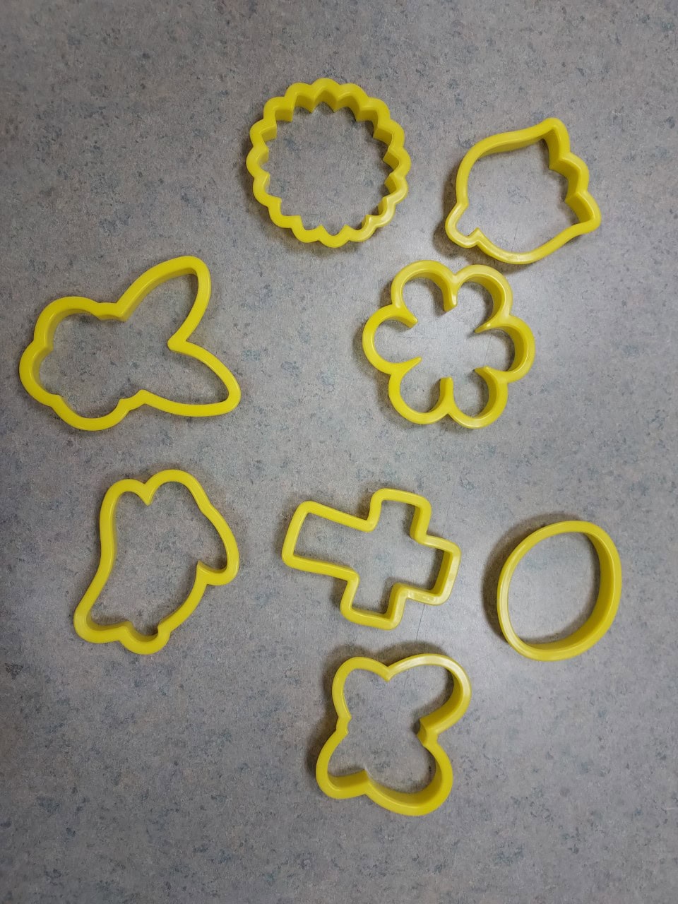Eight yellow plastic cookie cutters in various shapes—including a bunny, flower, egg, chick, cross, butterfly, and two others—are arranged on a light gray speckled countertop.