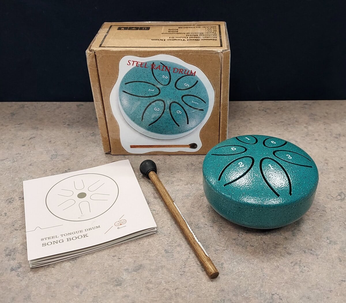 A small teal steel tongue drum, a wooden mallet, a song book, and the original cardboard box with a drum image on it are displayed on a gray surface against a dark background.