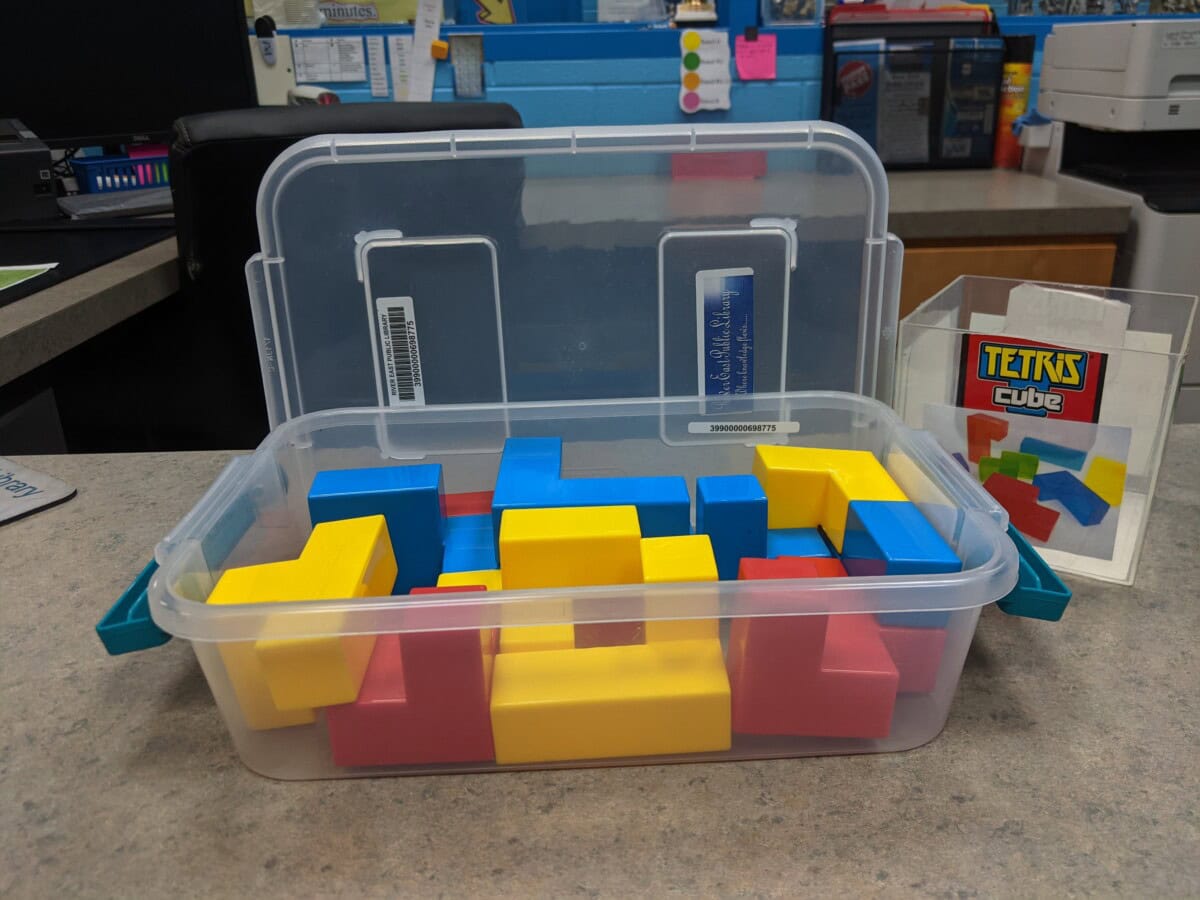 A clear plastic bin holds colorful red, blue, and yellow block pieces resembling Tetris shapes. Next to it, a card titled Tetris Cube is displayed. The setting appears to be an office or library counter.