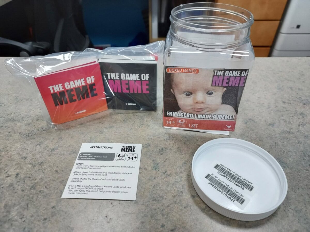 A card game set titled The Game of Meme on a countertop, with a clear jar, two card packs, an instruction card, and a lid. The jar label features a baby and text referencing internet memes.
