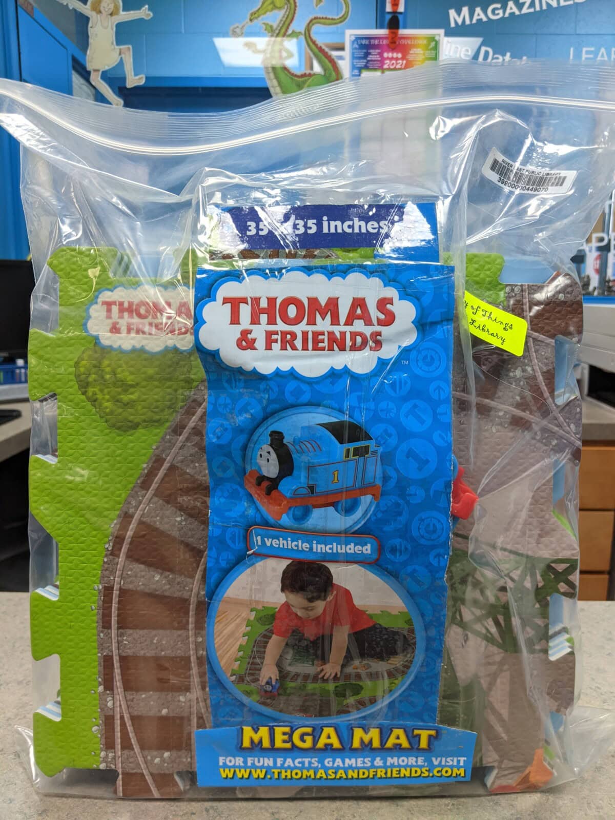 A sealed bag containing a Thomas & Friends Mega Mat playset, showing the packaging with an image of a child playing on the mat and a blue Thomas the Tank Engine toy vehicle included. A price sticker is visible on the bag.