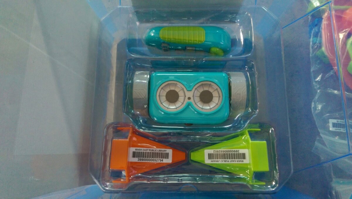 A plastic package containing a blue and green toy car, an orange and green ramp, and a small blue and green cutting tool, all organized in separate compartments.