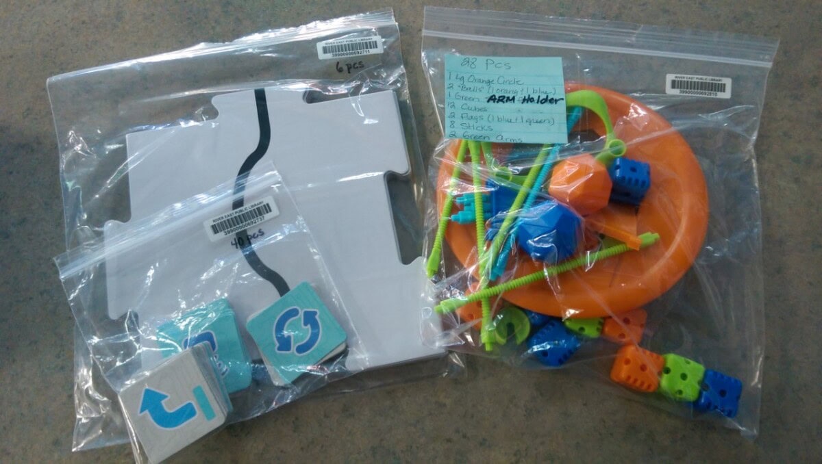 Clear plastic bags containing various learning materials: puzzle pieces, square tiles with arrows, colorful plastic rings, lacing strings, blocks, and a handwritten list labeling the contents. All items are on a tabletop.