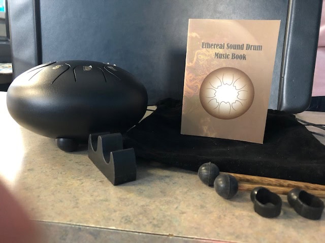 A black ethereal sound drum, mallets, and rubber feet rest on a table next to a booklet titled “Ethereal Sound Drum Music Book,” all placed on a black cloth.