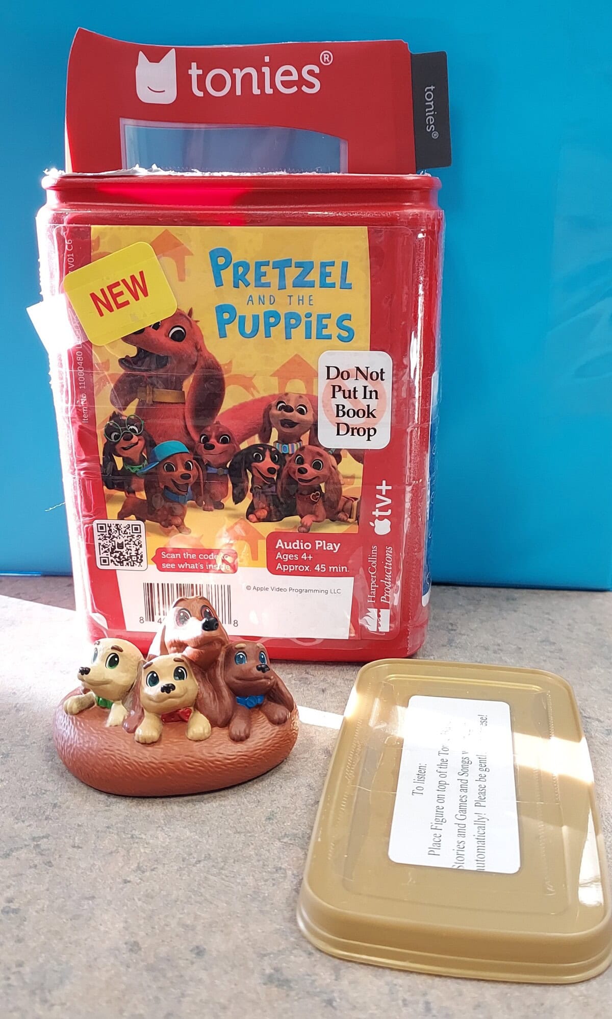 A Tonies audio play set titled Pretzel and the Puppies features a red box with cartoon puppies on the cover, a small figurine of three puppies, and a tan plastic case, all placed on a tabletop.