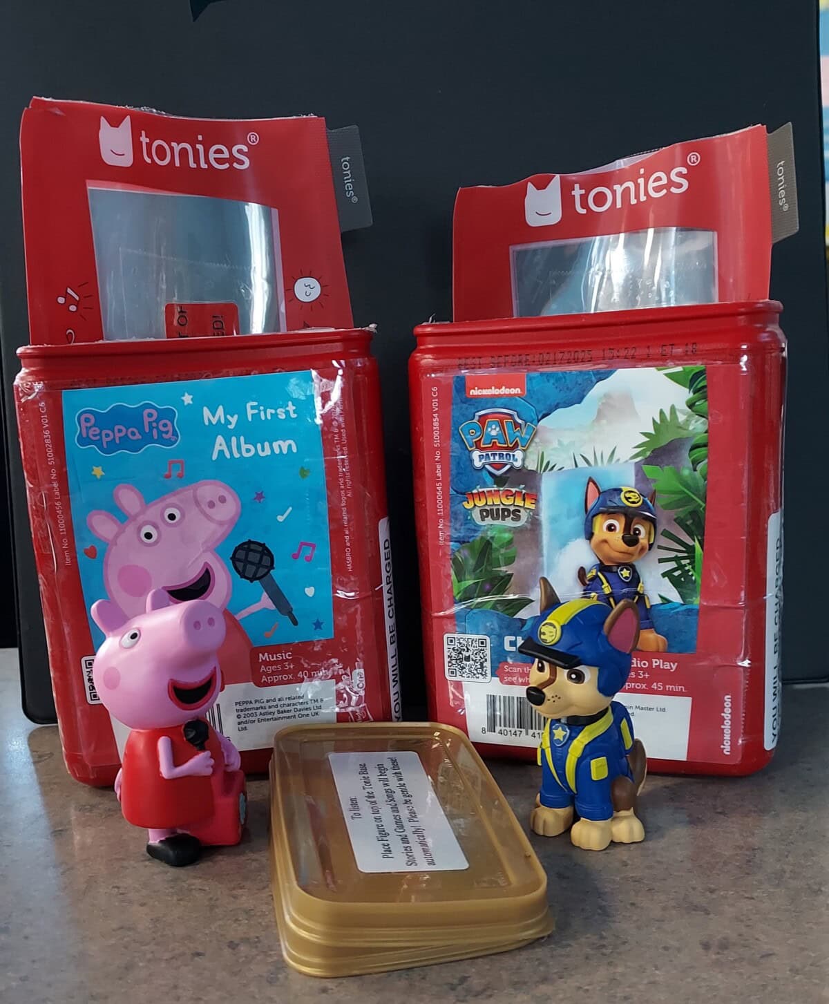 Two red Tonies boxes for Peppa Pig and PAW Patrol stand side by side. In front are Peppa Pig and Chase figurines, plus a small rectangular container with a white label, all on a countertop.