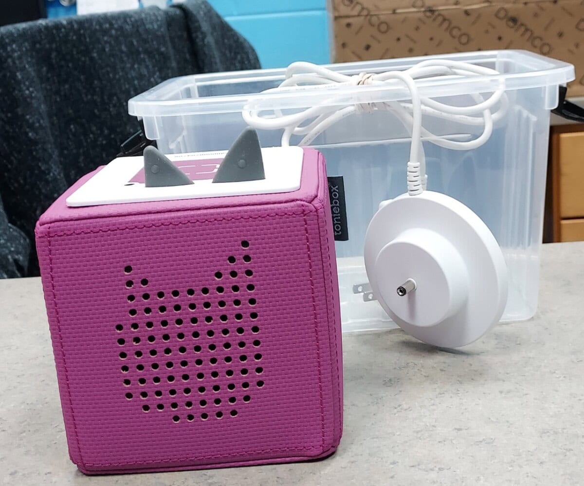 A pink Toniebox audio player with cat-shaped speaker holes and gray ears is placed on a gray surface. A white charging base and cord rest in a clear plastic storage container behind it.