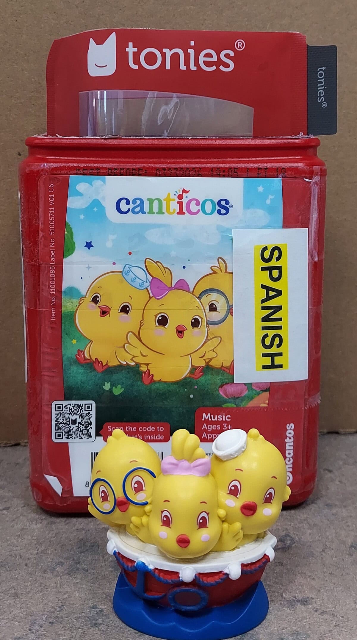 A red Tonies box labeled Canticos and Spanish with cartoon yellow chicks on the front sits behind a small figurine of three yellow chicks in a blue and red basket. One chick wears glasses.