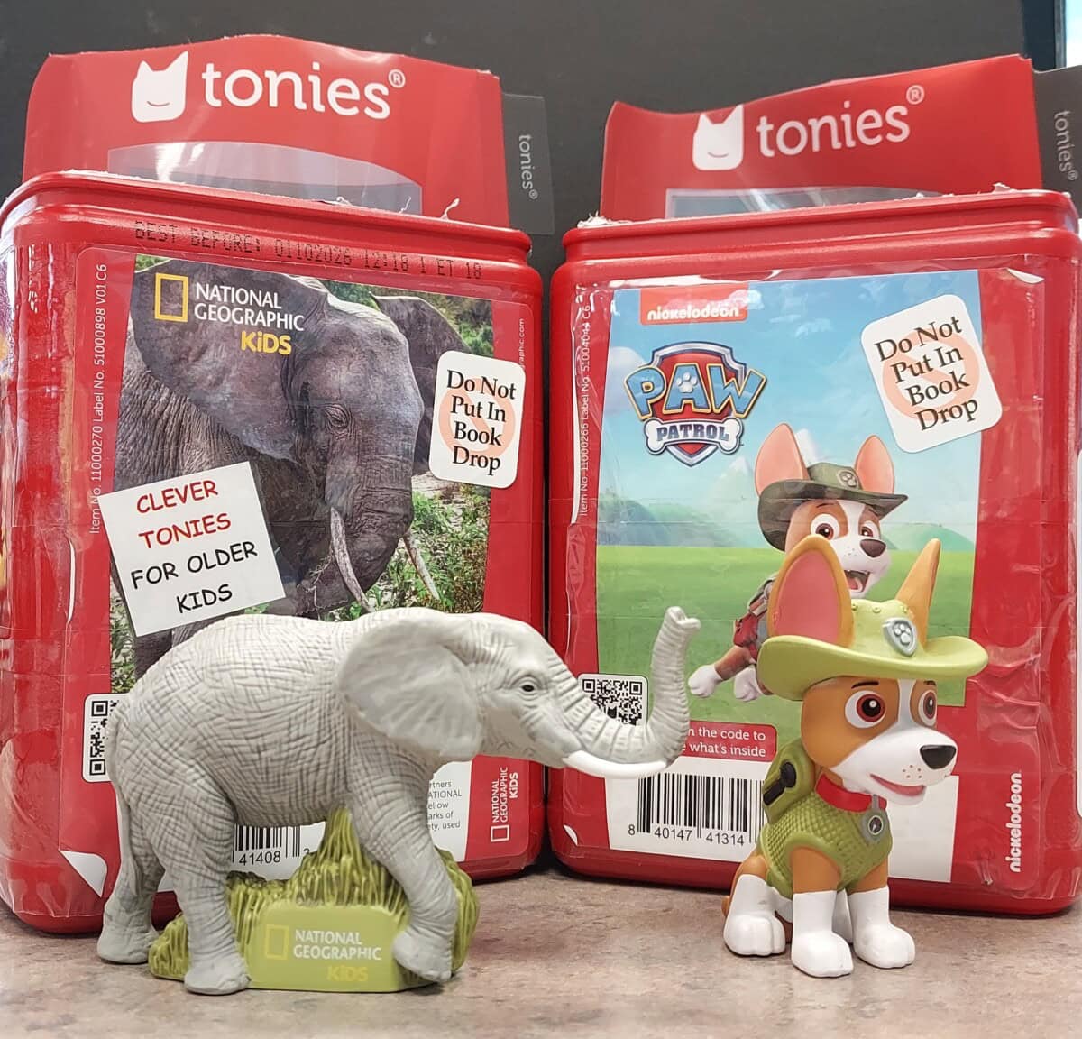Two red Tonie audio boxes with labels, one featuring National Geographic Kids with an elephant, and the other Paw Patrol with a dog figurine. Toy figures of an elephant and a Paw Patrol dog stand in front.