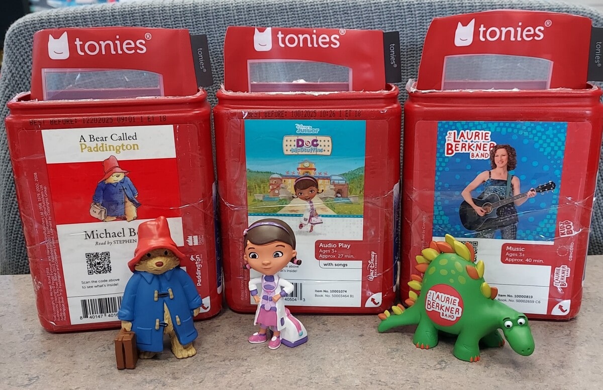 Three red Tonie audio boxes sit in a row, each featuring a different character: Paddington Bear, Doc McStuffins, and the Laurie Berkner Band, with matching character figurines standing in front of each box.
