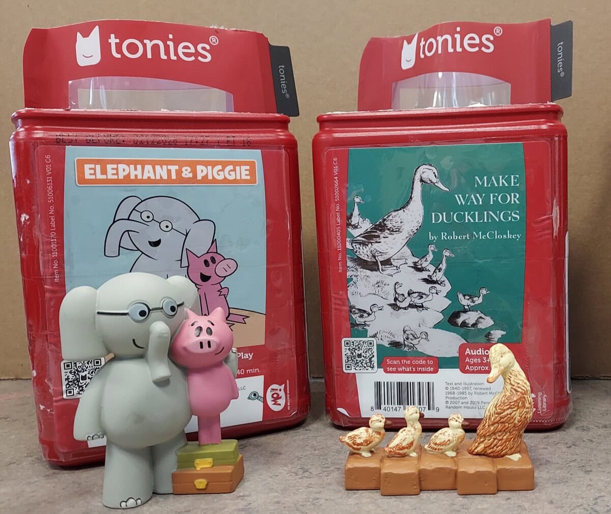 Two Toniebox audio figures—Elephant & Piggie and Make Way for Ducklings—stand in front of matching red Tonie packages with illustrated covers showing the respective book characters.