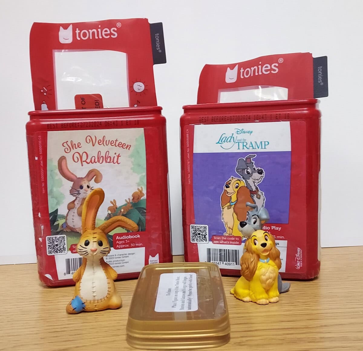 Two red Tonies audio boxes stand side by side. The left box features The Velveteen Rabbit and a rabbit figure; the right, Lady and the Tramp with dog figures of Lady and Tramp. A small brown tin sits in front.
