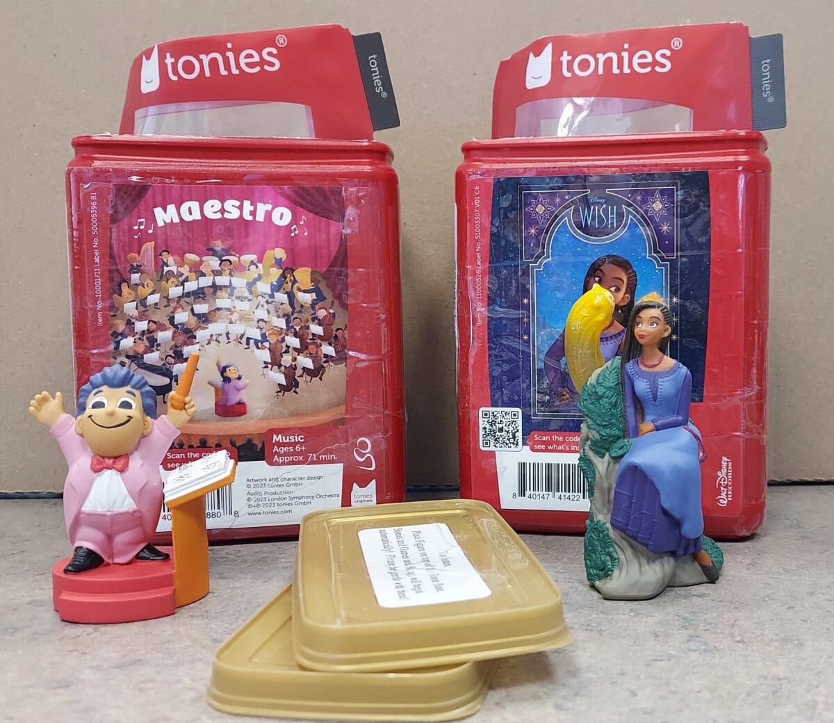 Two Tonies audio figures are in front of their packages: one is a conductor in a pink suit, and the other features two women sitting together. Both packages are red and upright, with labels and images on them.
