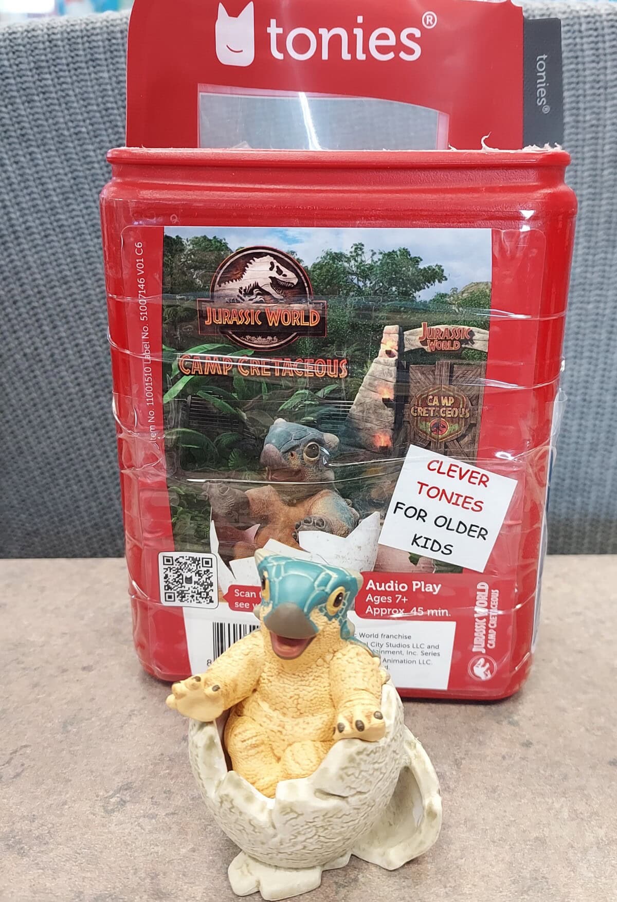A Tonies audio play box featuring Jurassic World: Camp Cretaceous is shown, with a small dinosaur figurine sitting in a cracked egg in front of the box. A label reads: “Clever Tonies for older kids.”.