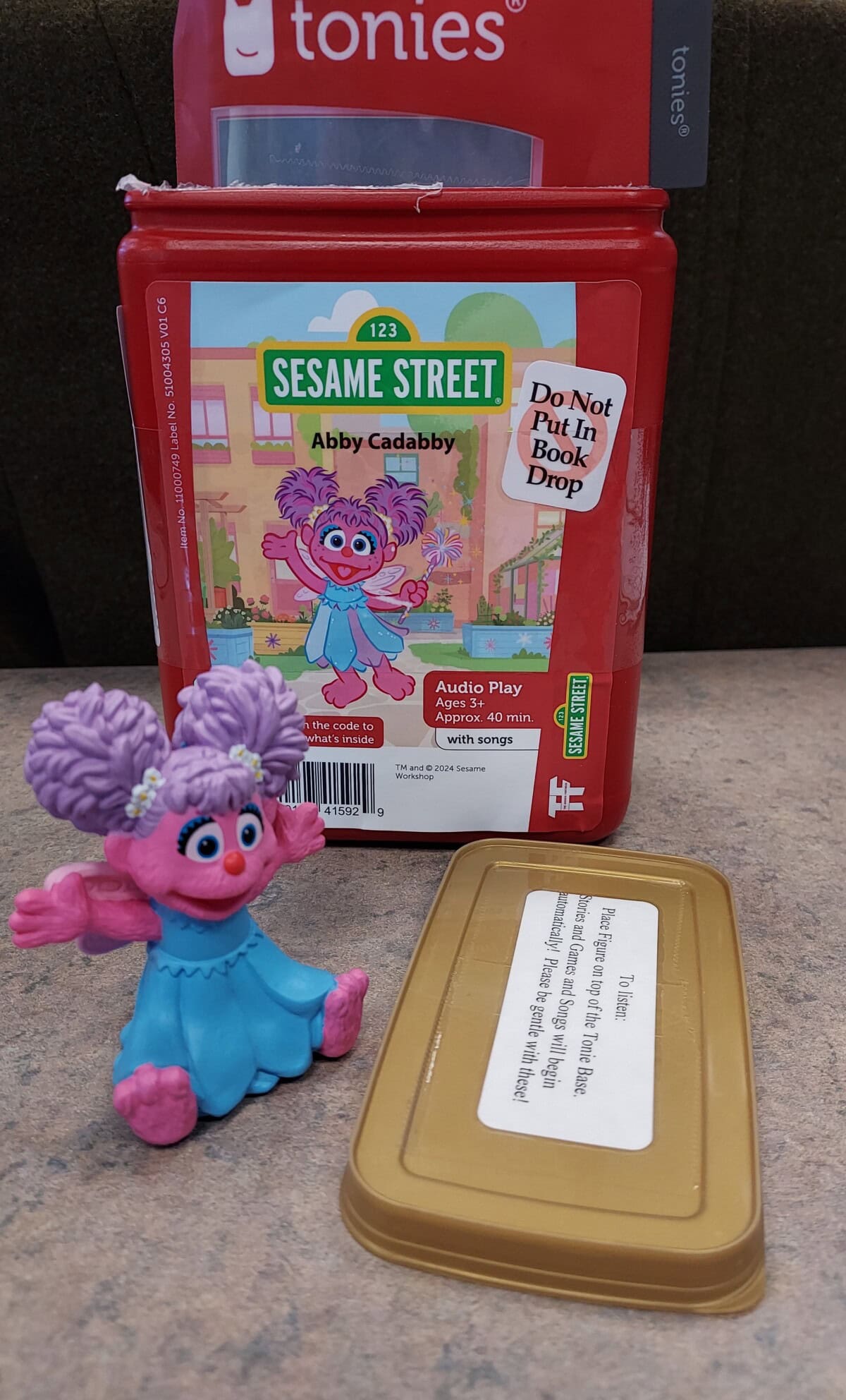 A small Abby Cadabby figurine sits in front of a Sesame Street Tonies audio play case labeled Abby Cadabby. The case lid is off and lying flat on the table nearby.