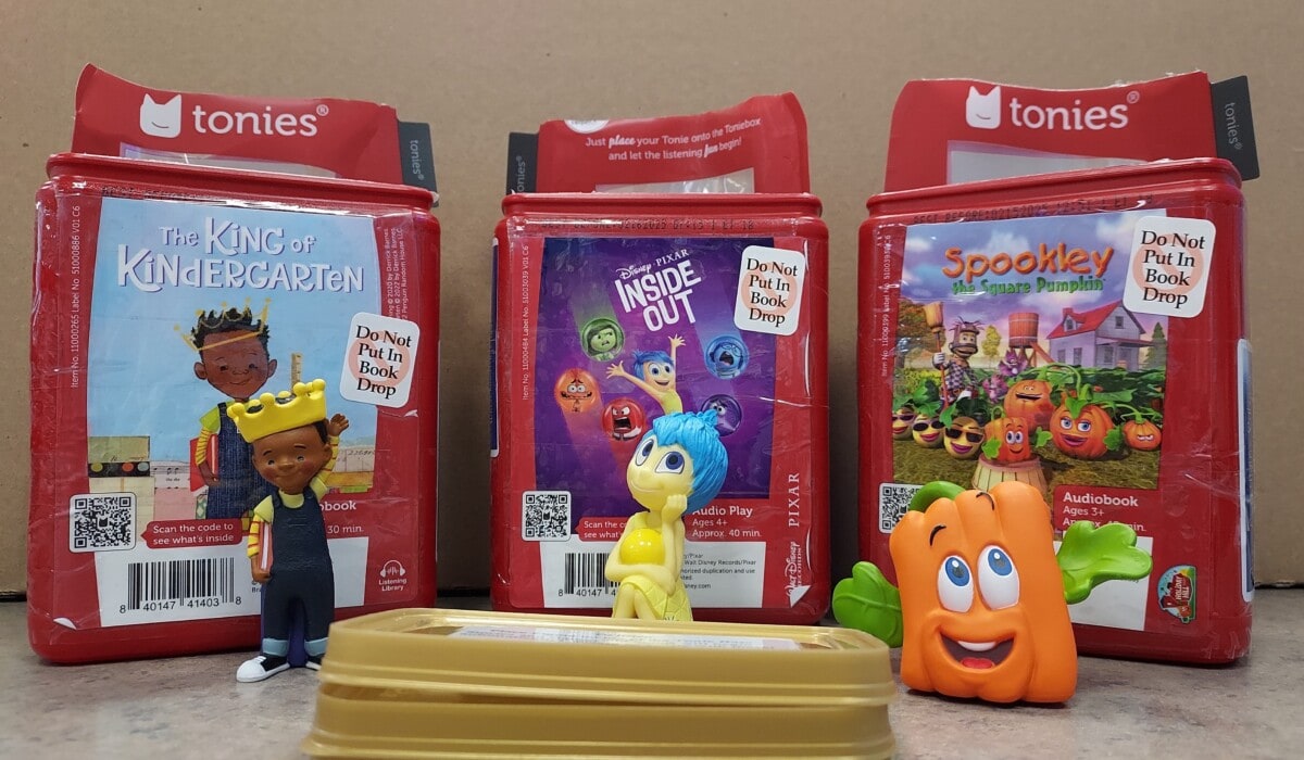Three children’s audiobook cases with Do Not Put in Book Drop signs stand upright. In front, toy figures from each story are displayed: a boy with a crown, Joy from Inside Out, and a smiling pumpkin.