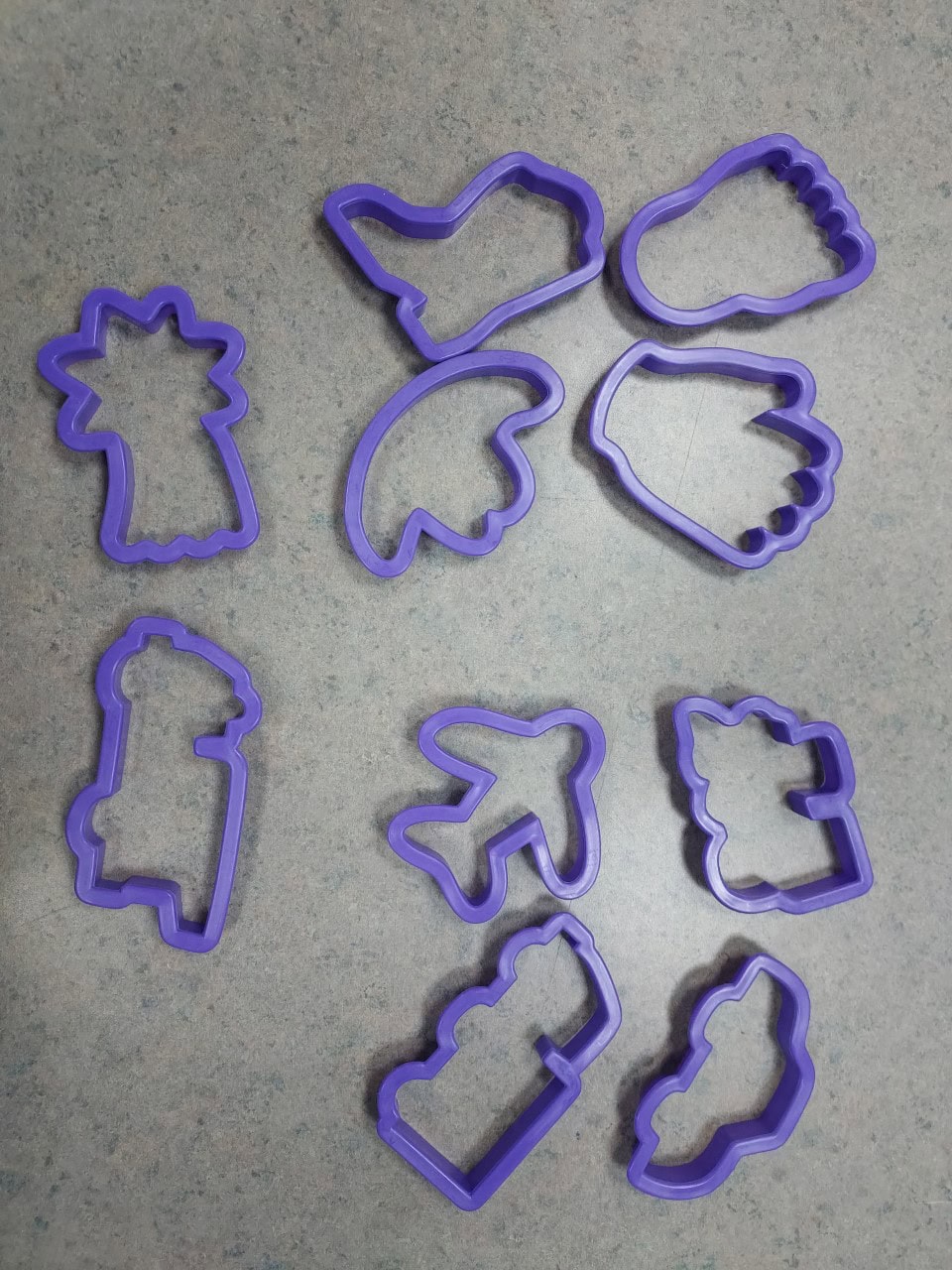 Nine purple dinosaur-shaped cookie cutters are arranged on a speckled gray countertop, each cutter forming a different dinosaur species or footprint. The cutters are evenly spaced in three columns.