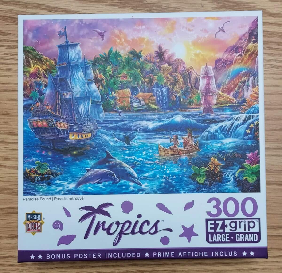 A colorful puzzle box displays a tropical paradise scene with a sailing ship, dolphins, a waterfall, lush vegetation, and two people rowing a boat. The box says Tropics, 300 EZ grip, and includes a bonus poster.