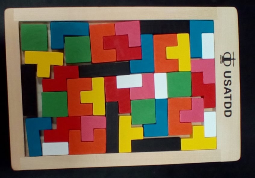 A wooden puzzle board labeled USATDD filled with colorful Tetris-like pieces in red, green, blue, yellow, pink, and white, all fitting together without gaps.