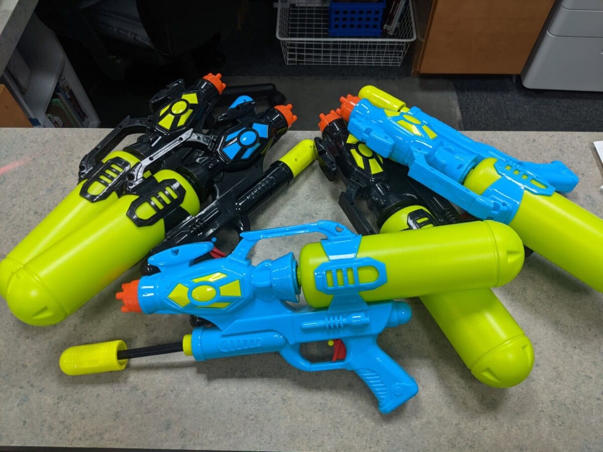 Four large, colorful water guns with yellow tanks and blue or black accents are placed on a countertop. Office supplies and shelving are visible in the background.